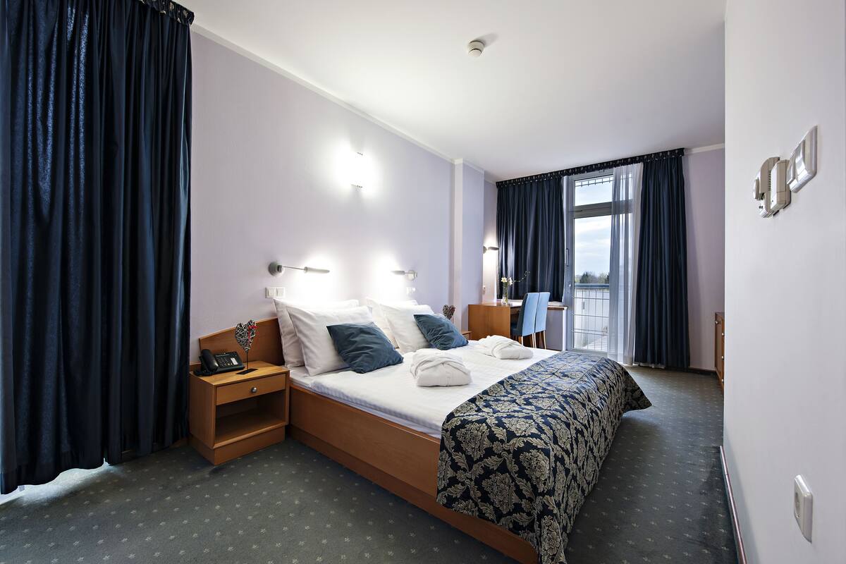 Habitación de hotel con cama, mesita de noche, lámpara y vistas al exterior a través de una ventana.