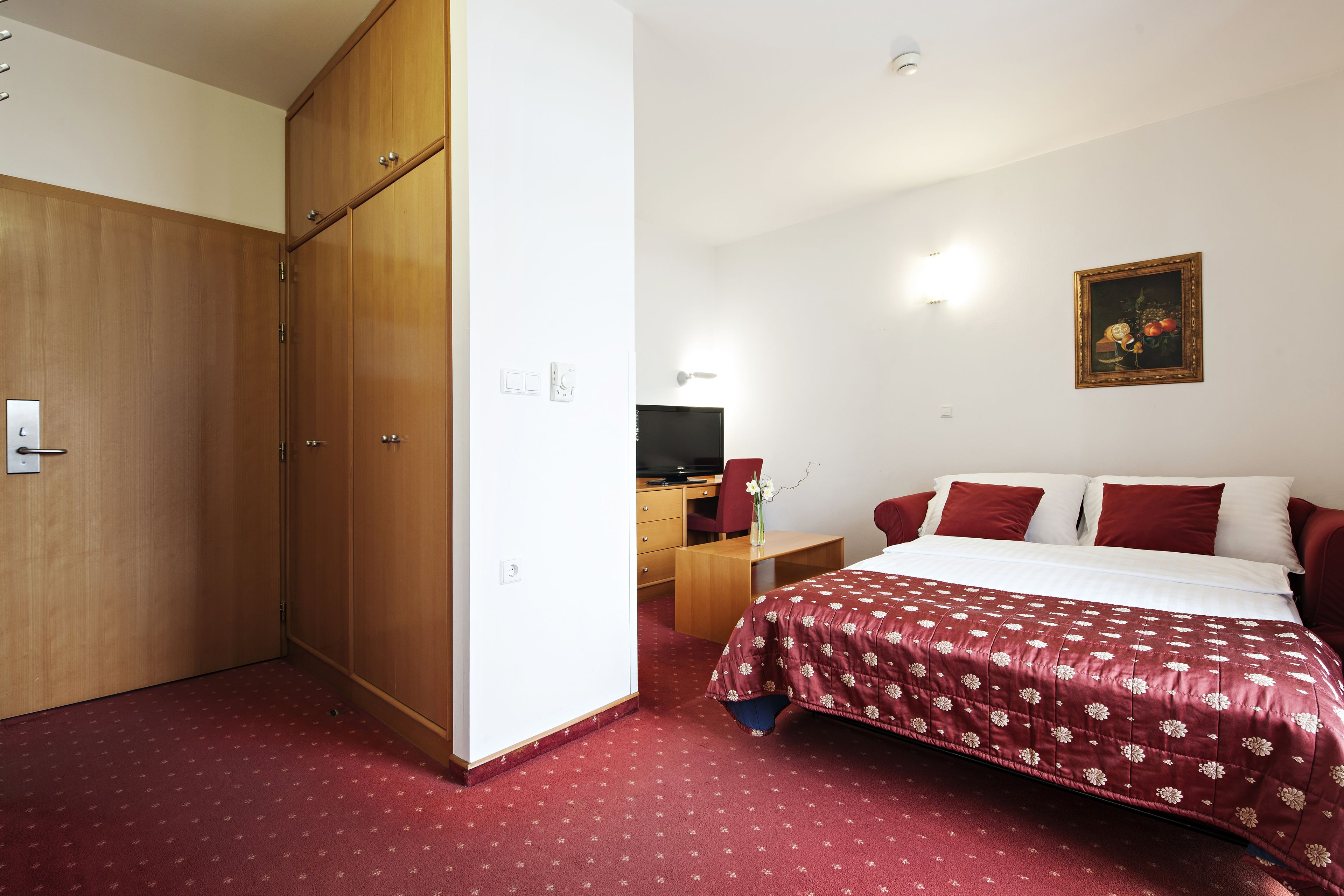 A hotel room with a large wardrobe, a bed with a red and white bedspread, a desk with a chair, and a television.