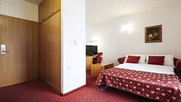 A hotel room with a large wardrobe, a bed with a red and white bedspread, a desk with a chair, and a television.