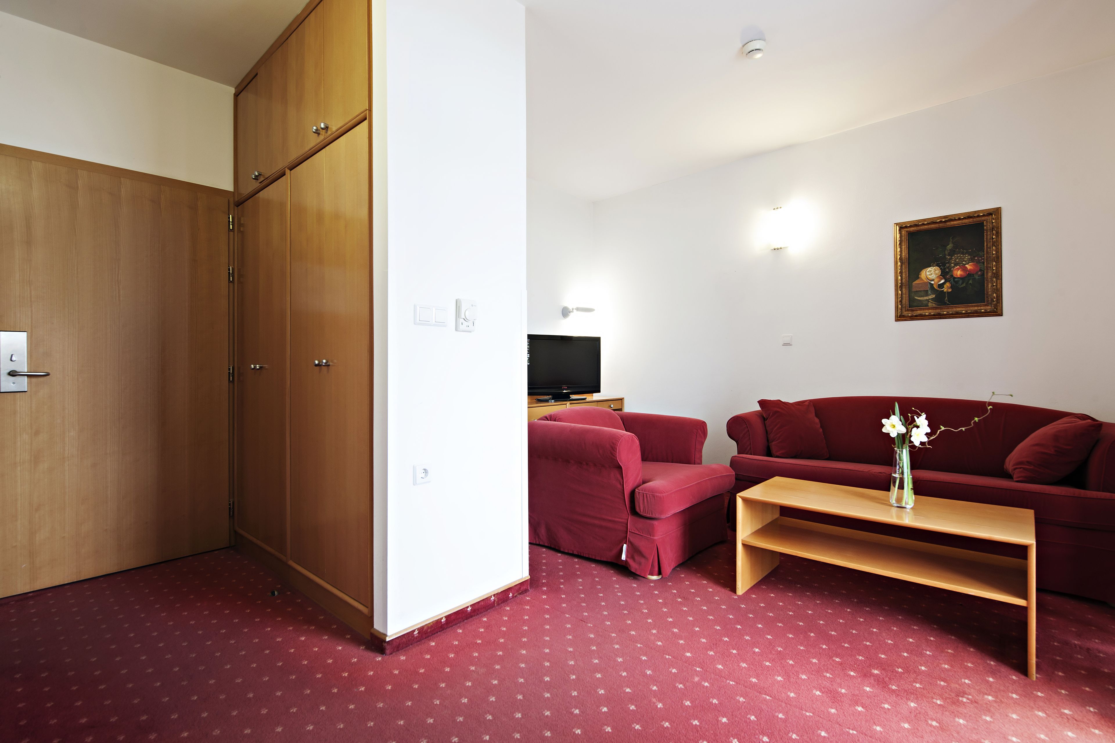 Een kamer met een houten garderobe, een rode bank, een houten salontafel en een schilderij aan de muur.