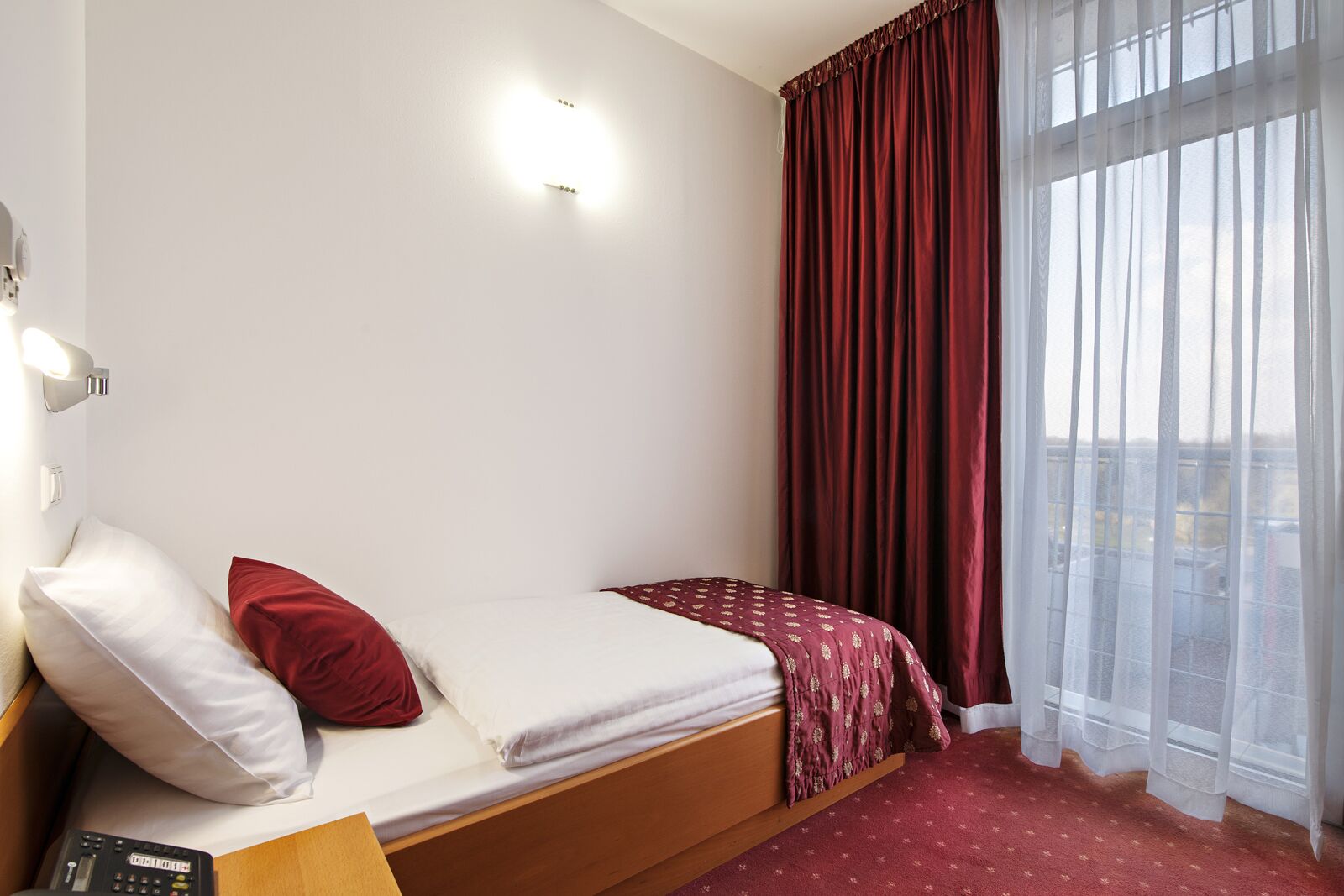 Una cama individual con colcha roja y blanca, almohada roja y cabecera de madera. Un control remoto se encuentra sobre una mesita de noche de madera junto a la cama. La habitación tiene un ventanal con cortinas blancas traslúcidas y vistas al exterior.