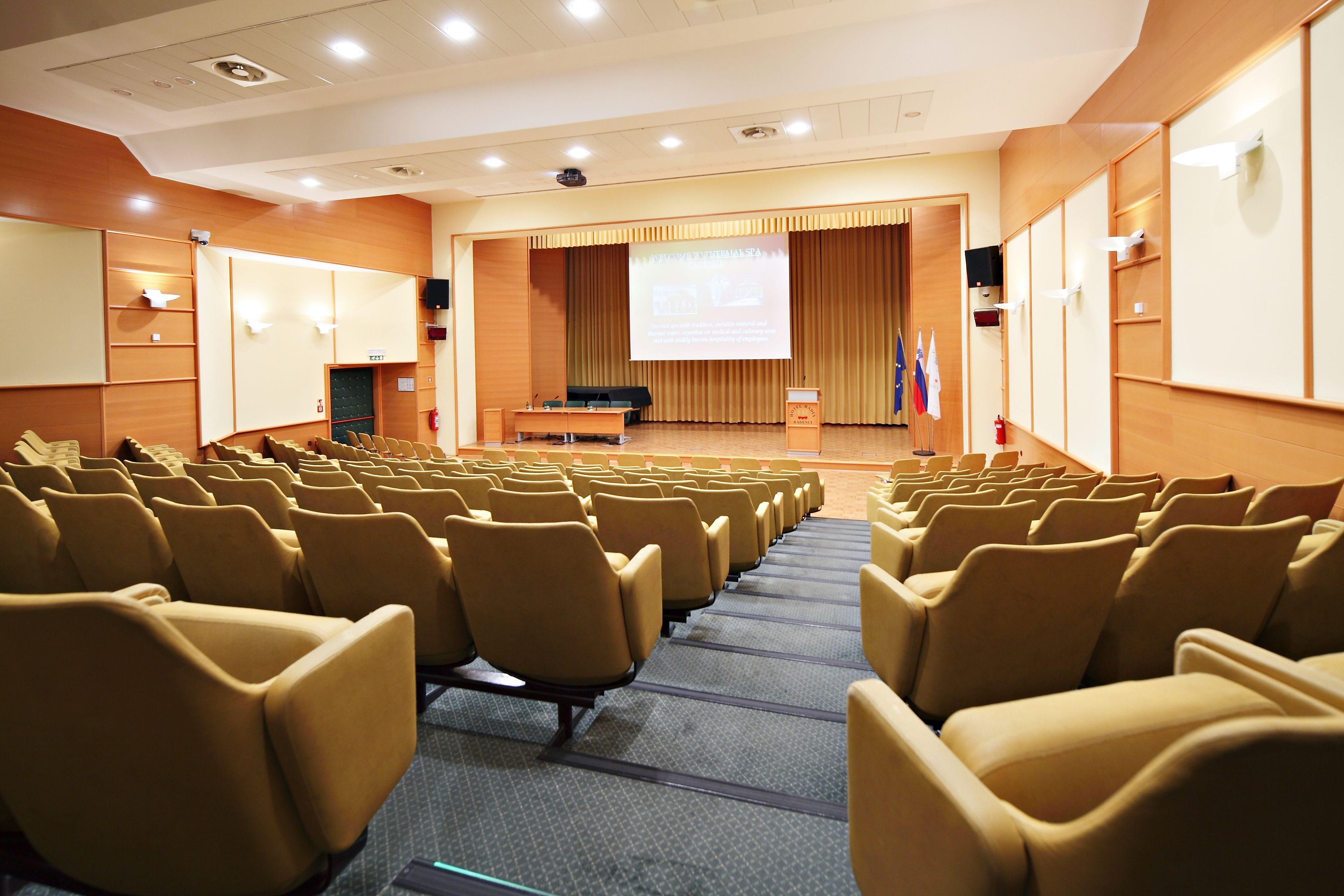 A conference room with rows of chairs, a podium, a screen, and flags.