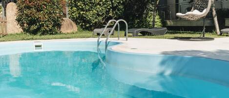 A swimming pool with a metal ladder, surrounded by greenery and a hanging chair.
