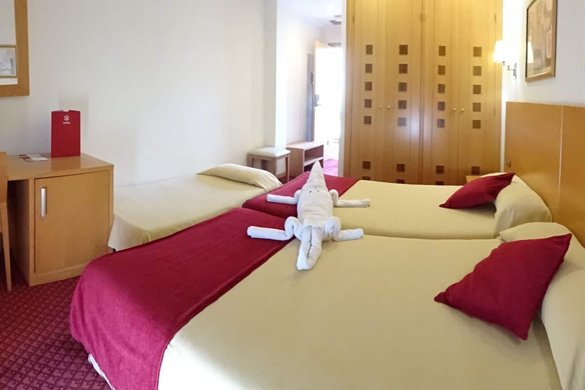 A hotel room with two beds, a wooden wardrobe, a desk, and a red suitcase.