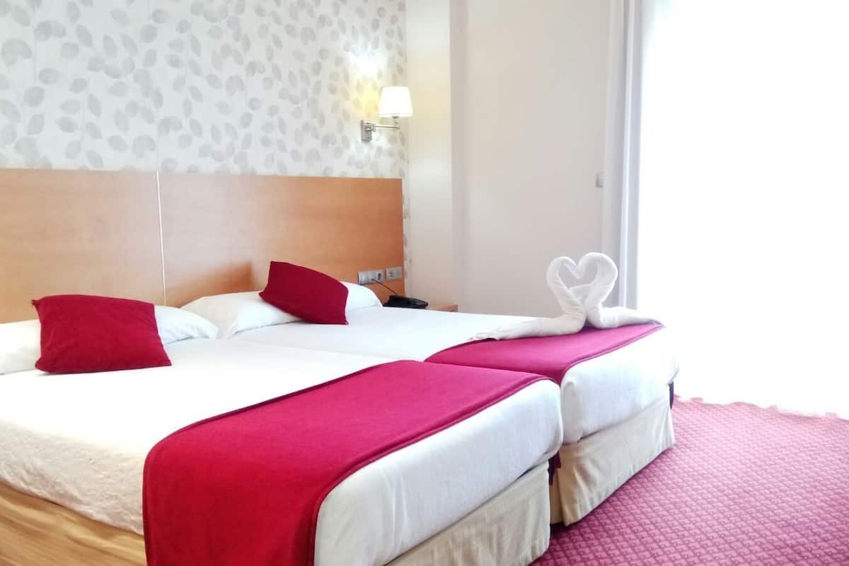 A hotel room with two beds, each with a red blanket and pillows, and a heart-shaped towel on the bed.