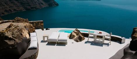 A sunlit outdoor lounge area with a pool, white lounge chairs, and a table with a red flower centerpiece, overlooking a calm sea and distant islands.