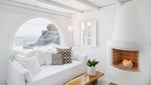 Une pièce blanche avec une grande fenêtre en arc de cercle, un canapé blanc orné de coussins à motifs, un banc en bois et une cheminée avec des bougies allumées.