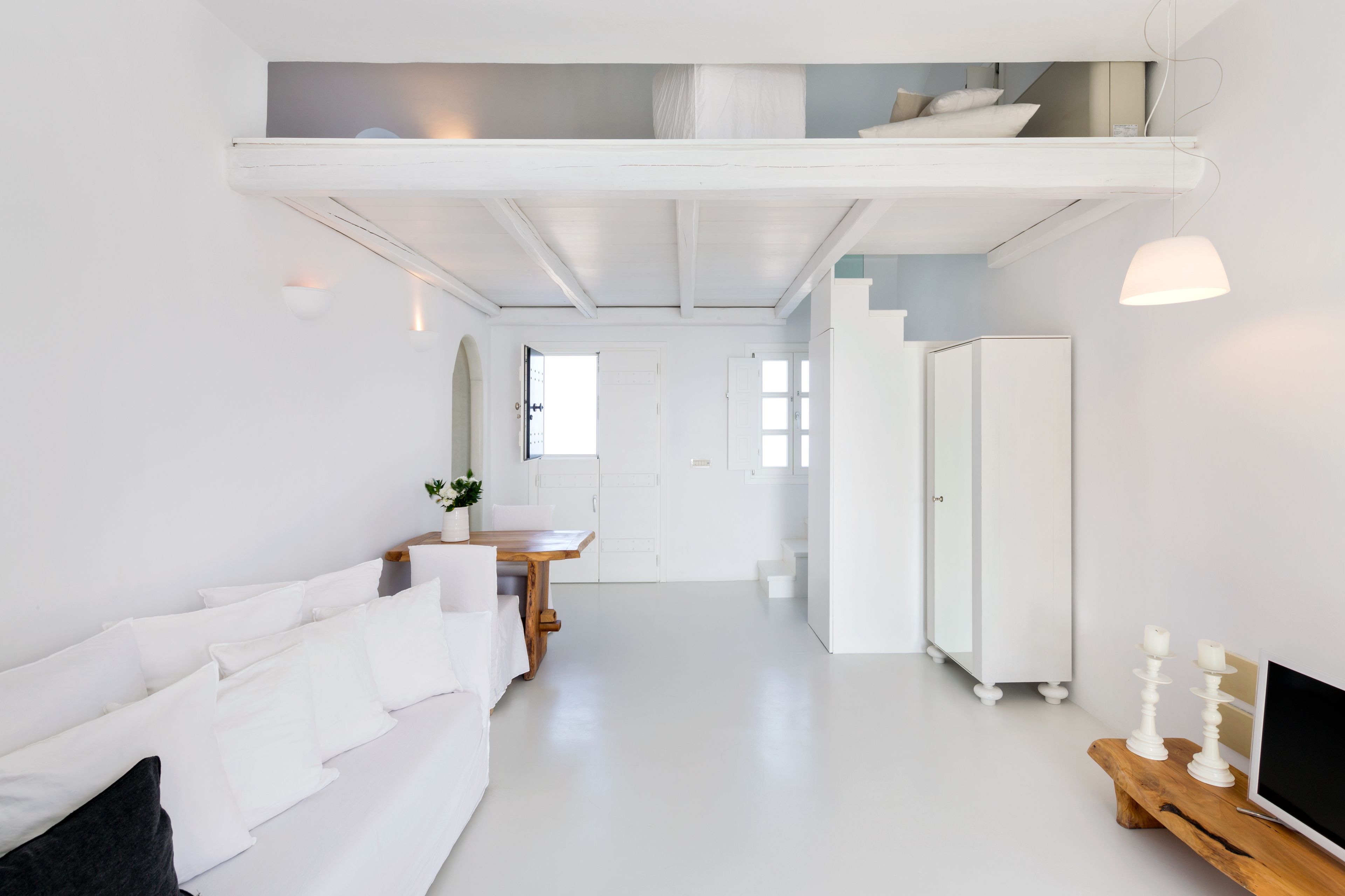 A modern, minimalist room with a white sofa, a wooden dining table, a TV on a wooden stand, and a wardrobe.