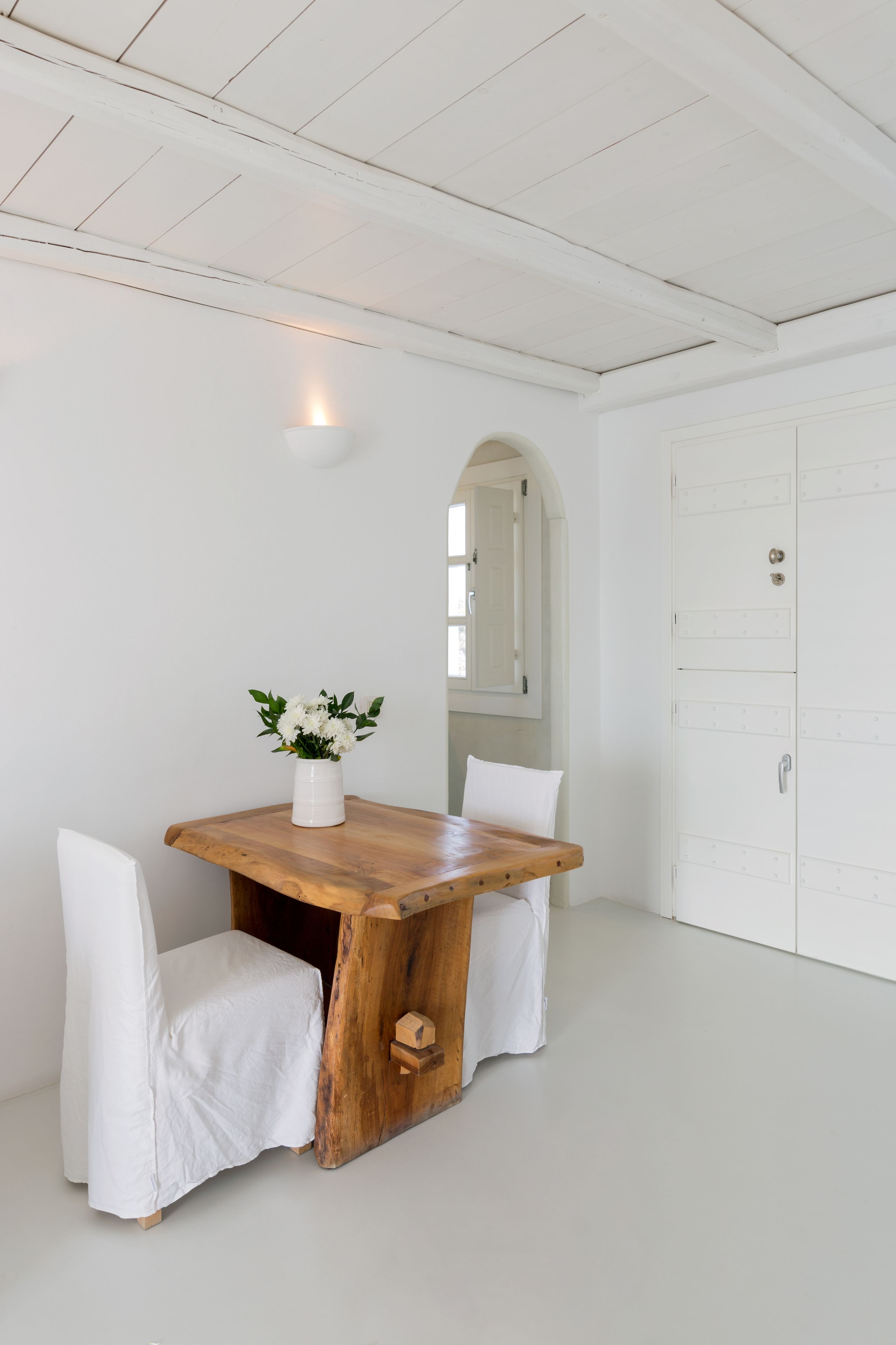 A white room with a wooden table, white chairs, a white vase with flowers, and a white wardrobe.