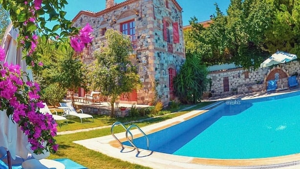 En byggnad av sten med en swimmingpool, omgiven av grönska och rosa blommor.