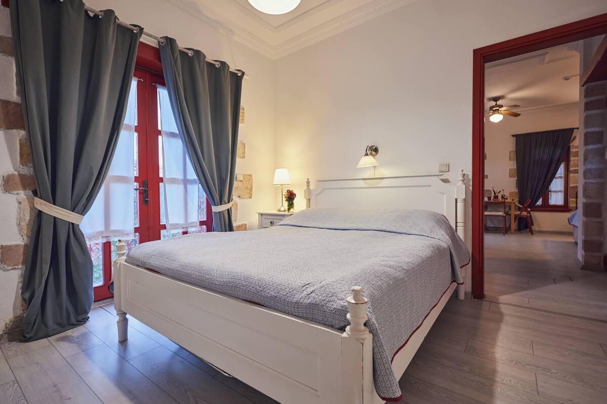 Ein Schlafzimmer mit einem weißen Bett, grauer Bettwäsche, einem Nachttisch mit Lampe sowie Blick auf eine rote Tür und ein Fenster.