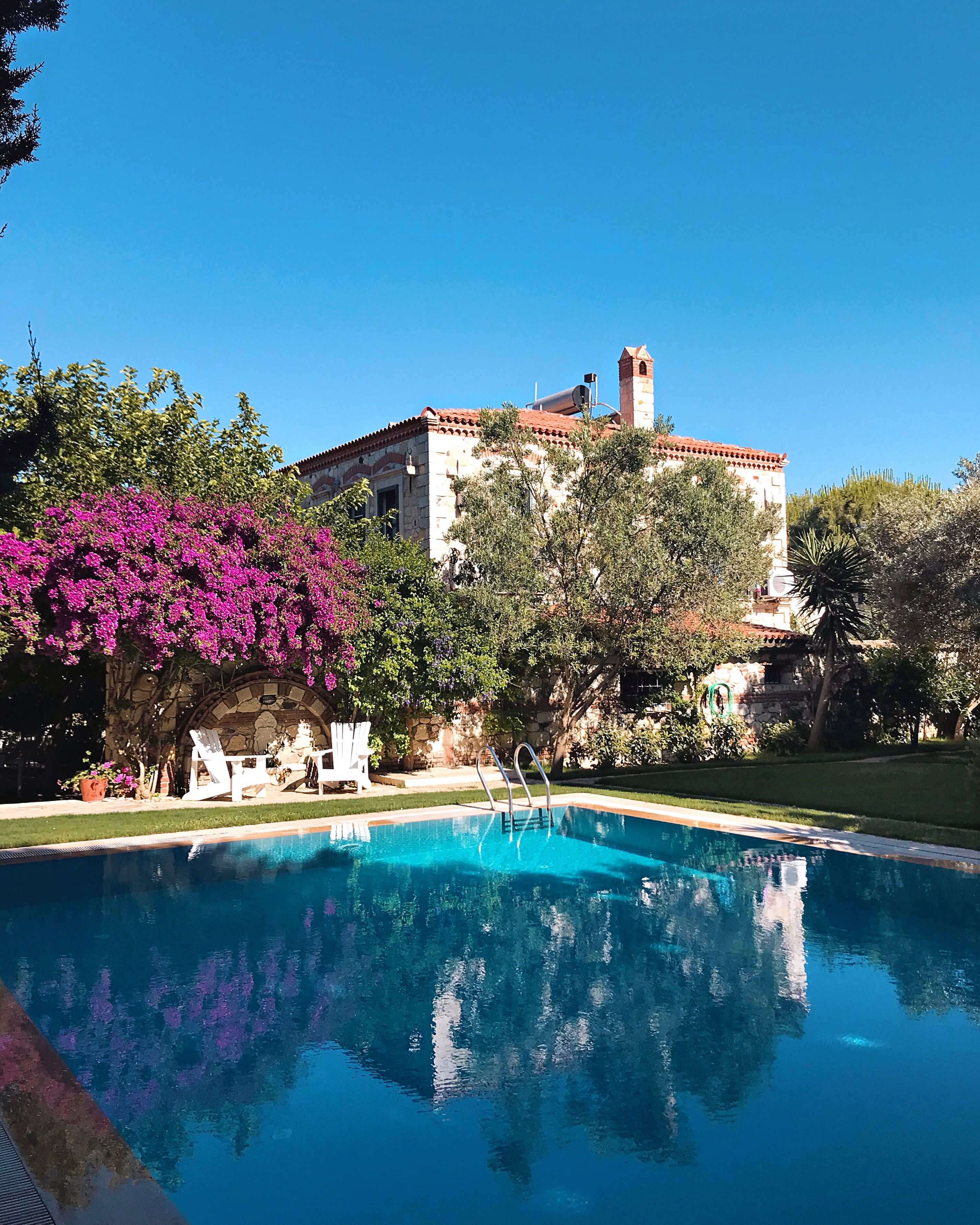 A swimming pool with a clear blue water surface, surrounded by lush greenery and a stone building with a tower.