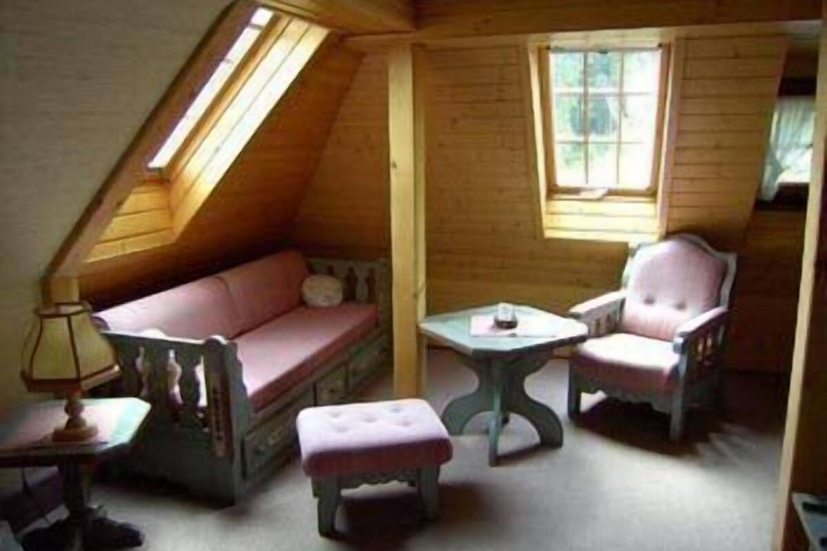 A cozy attic room with a sloped ceiling, a pink sofa, two matching chairs, a small table, and a lamp.