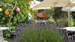 Un patio al aire libre con mobiliario de mimbre, una escultura de metal decorativa y plantas florecientes moradas.