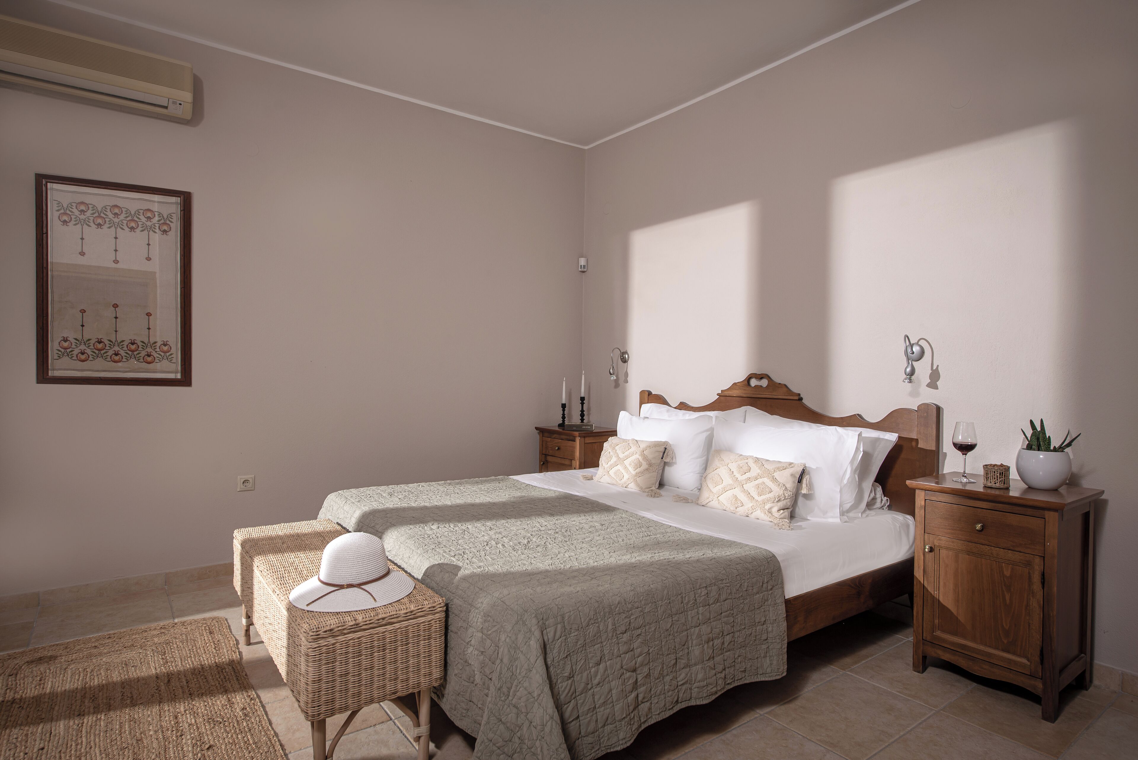 A bedroom with a large bed, two bedside tables, a nightstand, a wicker chair, and a wall-mounted air conditioner.
