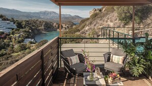 Un balcon avec deux chaises, une table ornée d’un vase et de fruits, et une vue sur un paysage côtier avec des bâtiments et des montagnes.