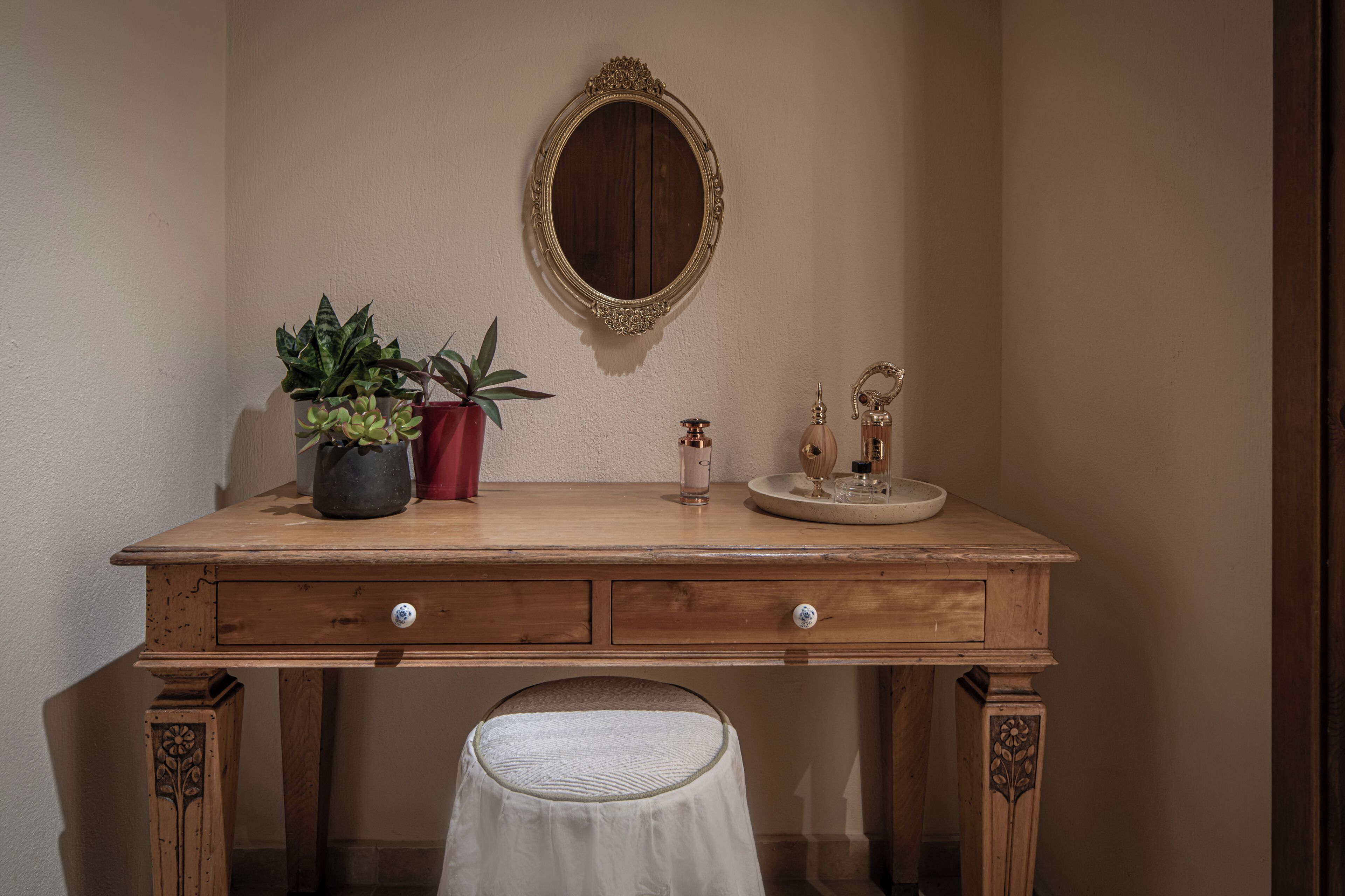 Un tocador de madera con un espejo, una planta en maceta y una mesita con un frasco de perfume y un vaso.