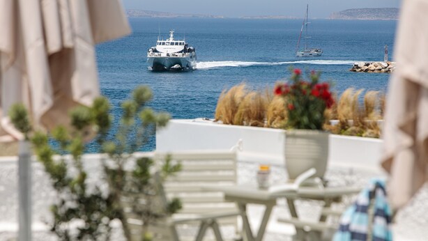 A boat on the water, a rocky outcrop, and a white building with a table and chairs in the foreground. - Lygdamis Hotel (Naxos)