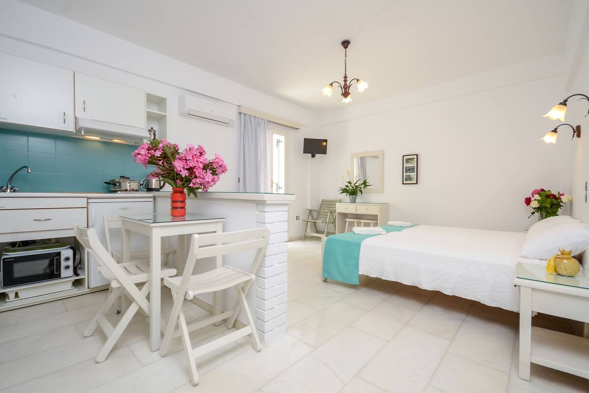 A modern, well-lit room with a kitchenette, a dining area with white furniture, a bed with a teal bedspread, and decorative flowers.