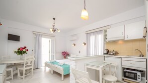 A modern, white-themed room with a bed, a dining table, a kitchen area, and a TV mounted on the wall. - Lygdamis Hotel (Naxos)