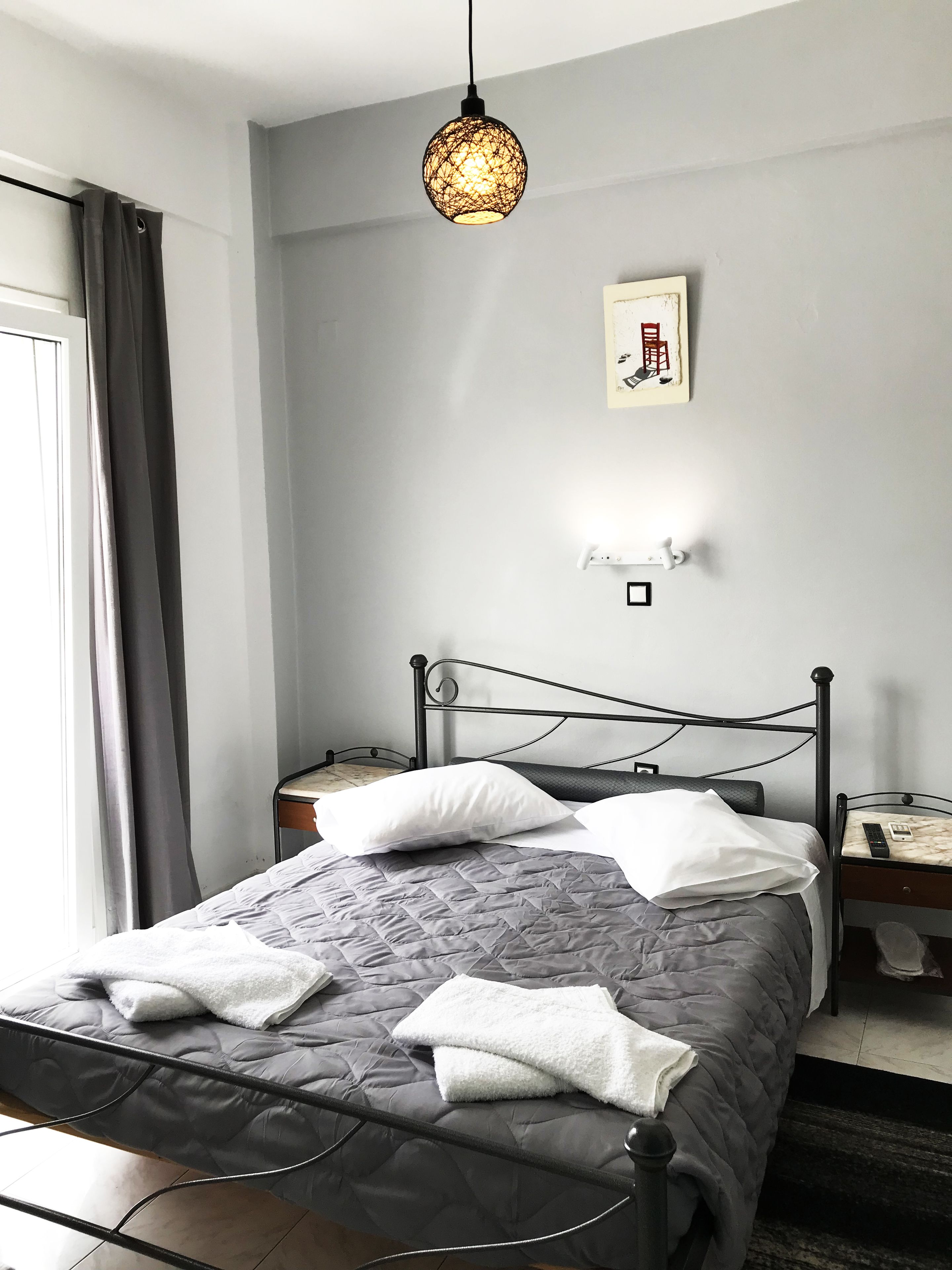 Cama bem arrumada com colcha cinza, travesseiros brancos e toalha branca. Cabeceira de metal preta e mesa de cabeceira com controle remoto são visíveis. Luminária pendente sobre a cama.