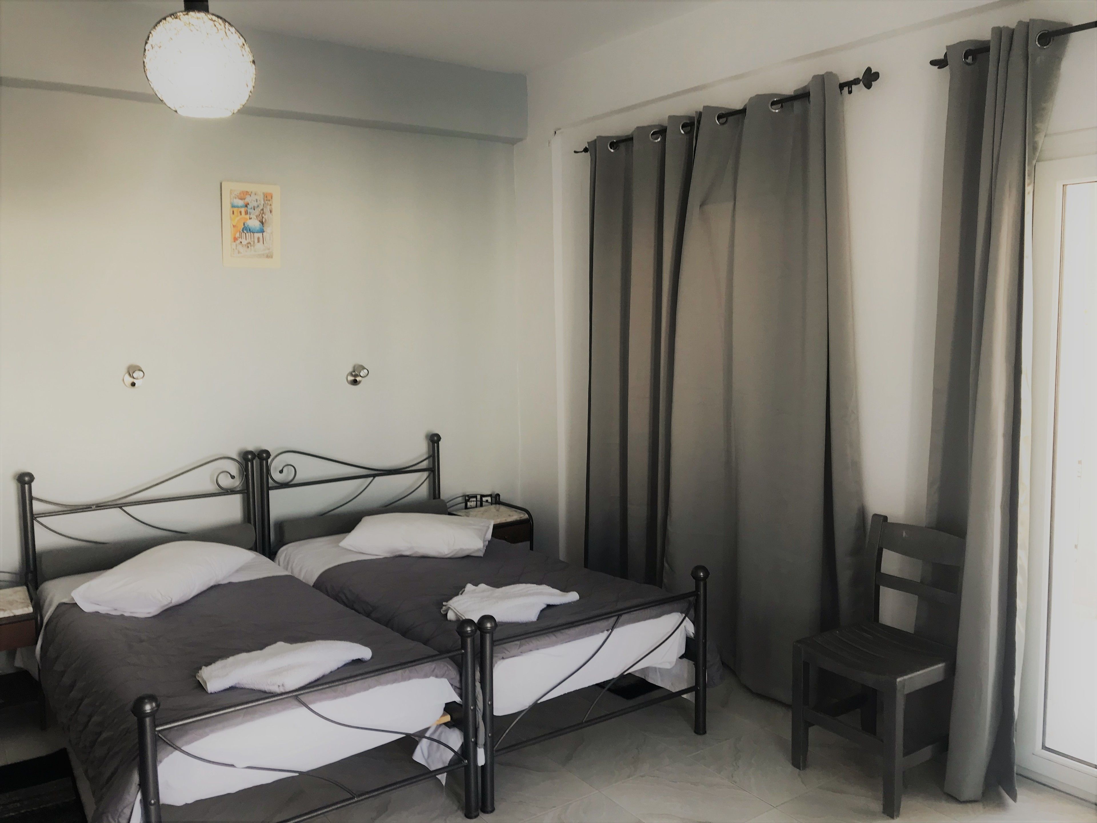 Une chambre à coucher avec un lit double, une chaise, une table de chevet, un luminaire suspendu et une fenêtre avec des rideaux.