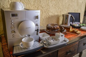 Una cafetera, una taza y un platillo, una bandeja con un pastel y cubiertos, y una tetera sobre una mesa.