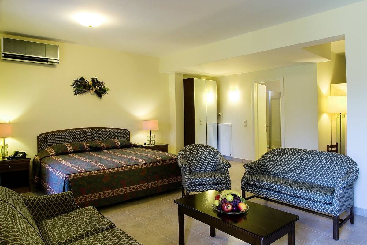 A hotel room with a bed, two armchairs, a coffee table with fruit, a mirror, and a radiator.