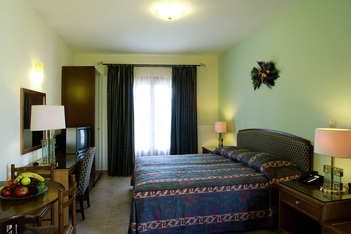 A hotel room with a bed, a desk with a computer, a television, and a dining table with fruit.