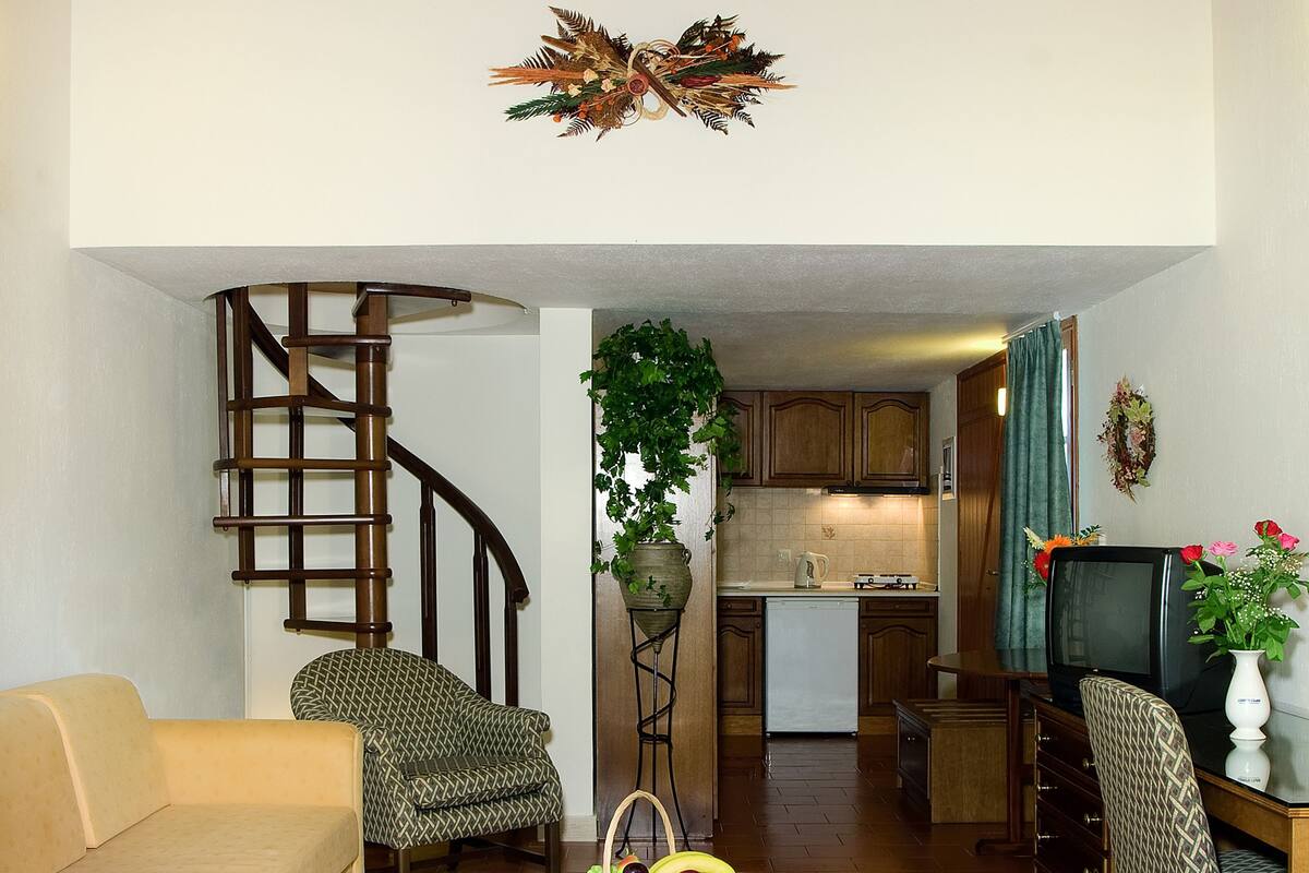 A living room with a sofa, armchair, and a coffee table with fruit. A spiral staircase leads to an upper level.