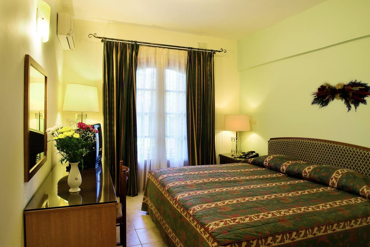 A hotel room with a bed, a desk with a lamp, a mirror, and a window with curtains.