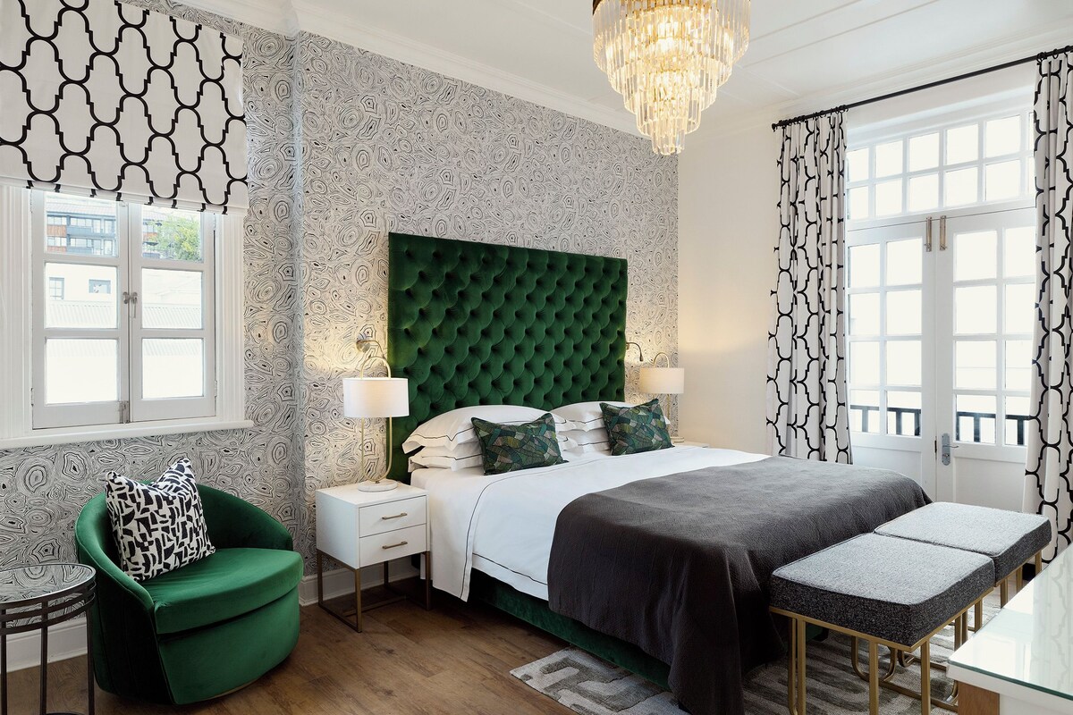 Een slaapkamer met een groen gewatteerd hoofdeinde, een nachtkastje met een lamp, een groene fauteuil en een bankje.