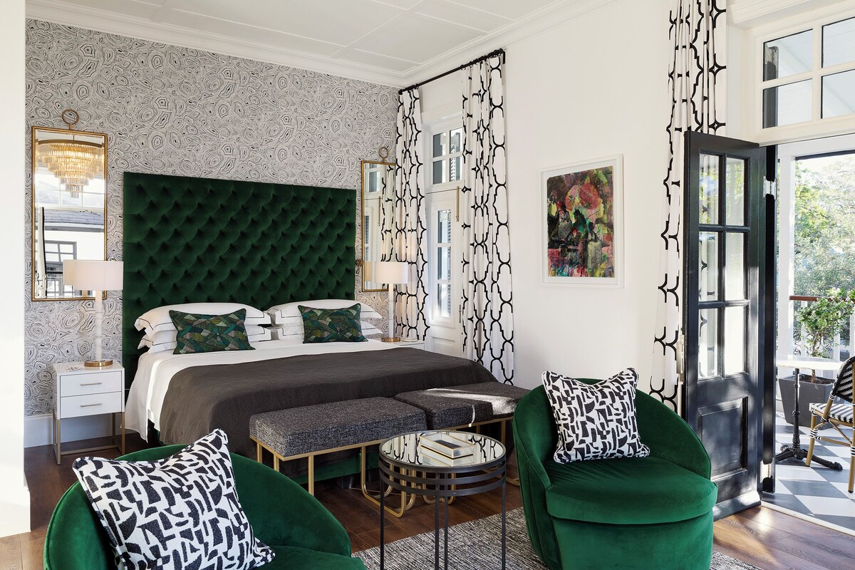Een slaapkamer met een gebonden hoofdeinde, een bed met gekleurde kussens, een bijzettafeltje, een groene fauteuil, en uitzicht op de omgeving via een deur.