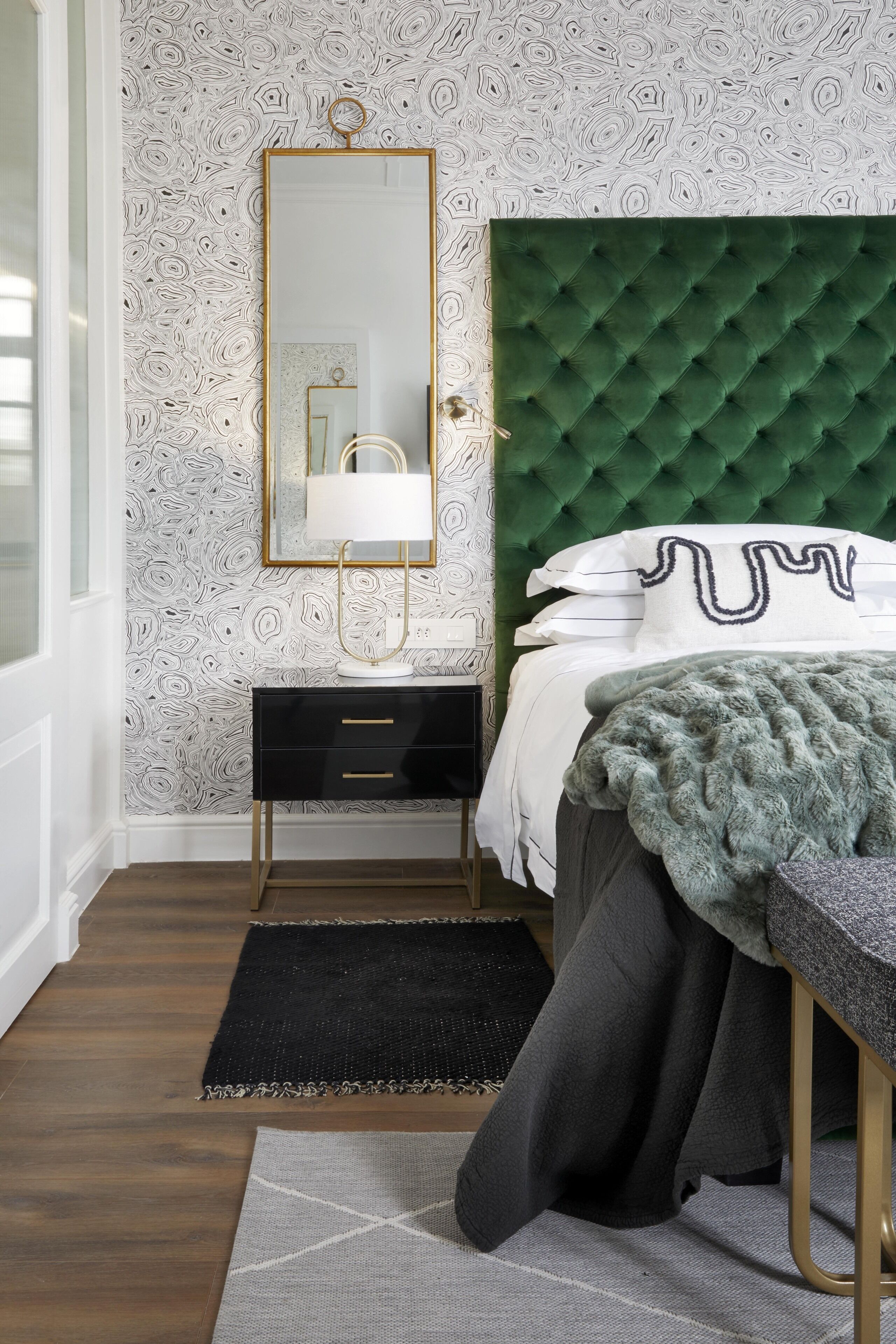 A bedroom with a green tufted headboard, a black bedside table, a gold-framed mirror, and a patterned wallpaper.