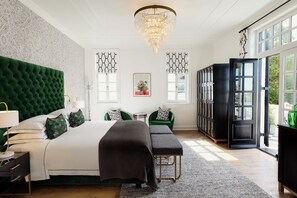 Een slaapkamer met een gestoffeerd hoofdeinde, een bed met kussens, een nachtkastje, een lamp, een bankje, een stoel, een klein tafeltje, een kroonluchter, een kast en een raam met uitzicht.