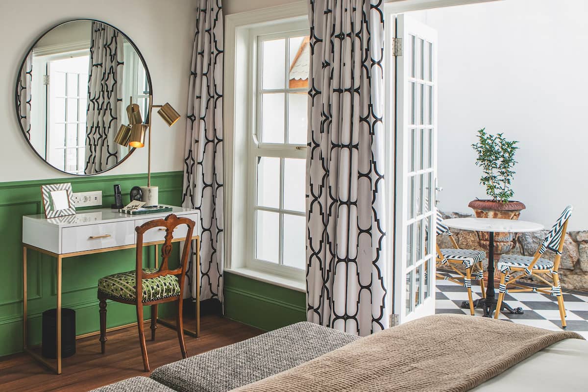 Een kamer met een groene wand, een ronde spiegel, een bureau met stoel, een bed met een bruine deken en een raam met patroon gordijnen.