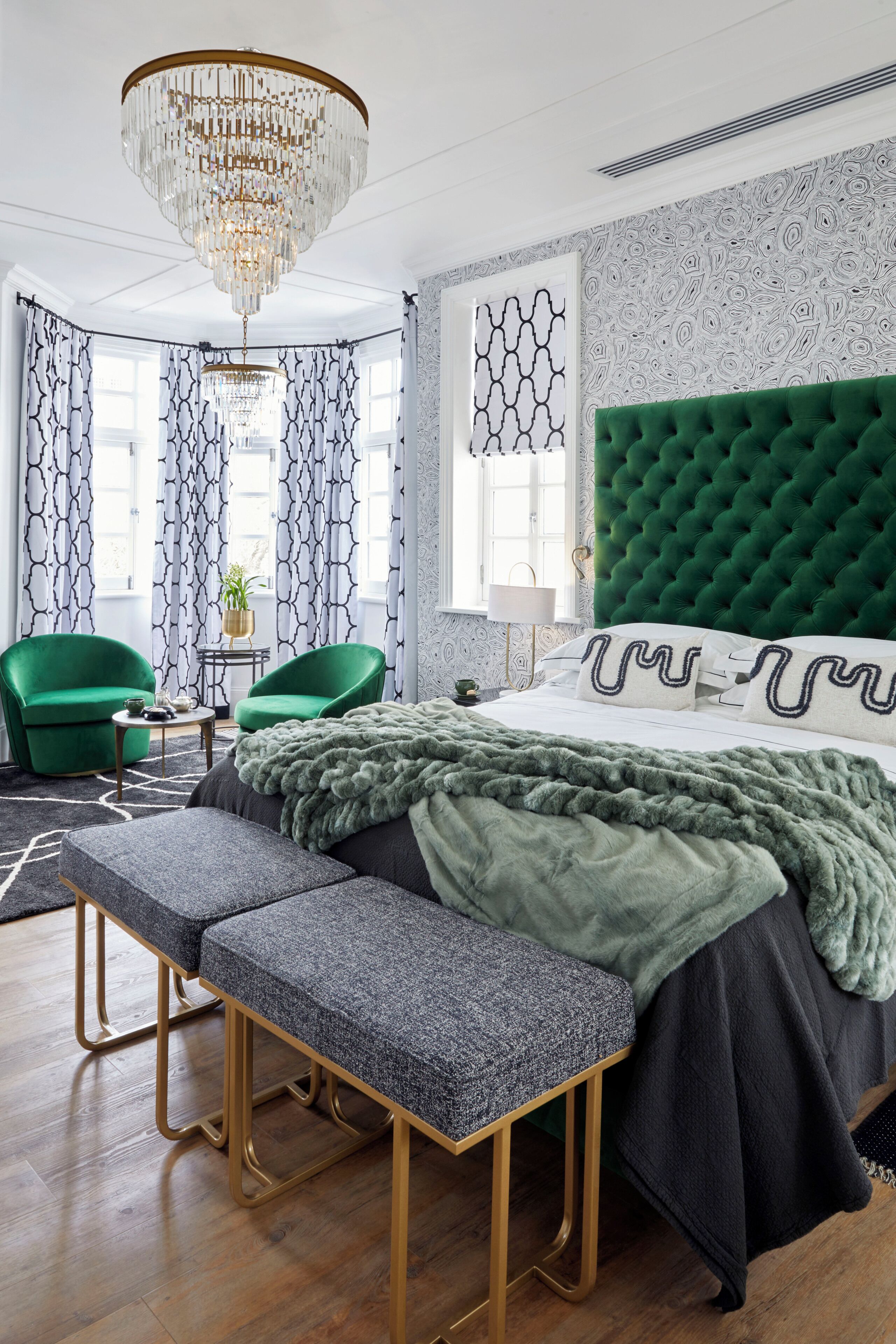 Un dormitorio con cabecera tapizada en verde, una cama grande con colcha de diseño gris y blanco, un banco al pie de la cama y una araña.