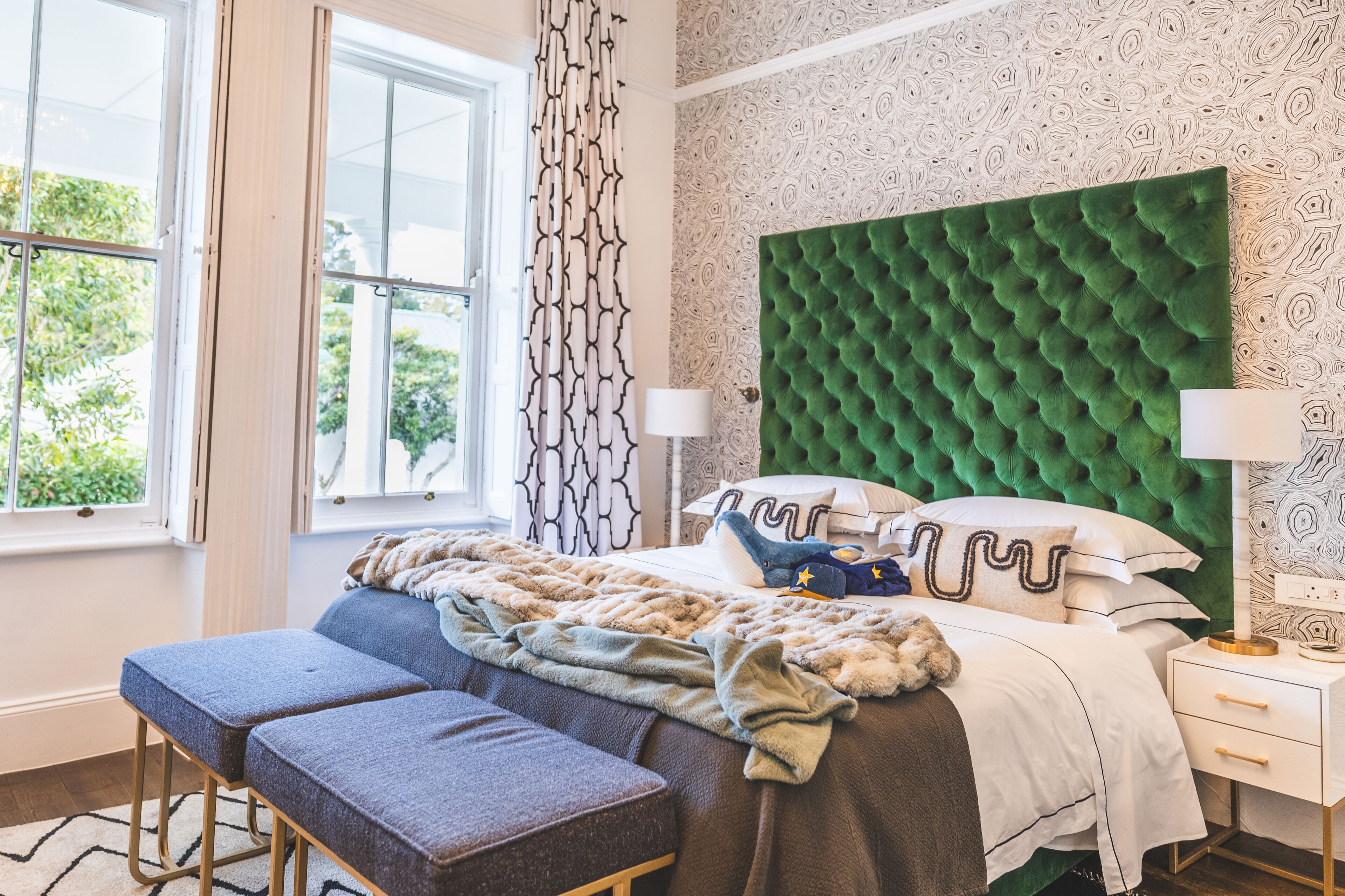 Quarto com cabeceira verde estofada, uma janela grande com cortinas brancas, mesa de cabeceira com luminária e um banco aos pés da cama.