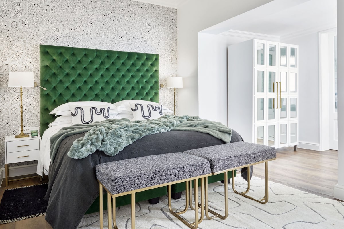 Een moderne slaapkamer met een groen gewatteerd hoofdeinde, een grijze bank en een witte garderobe met glazen deuren.