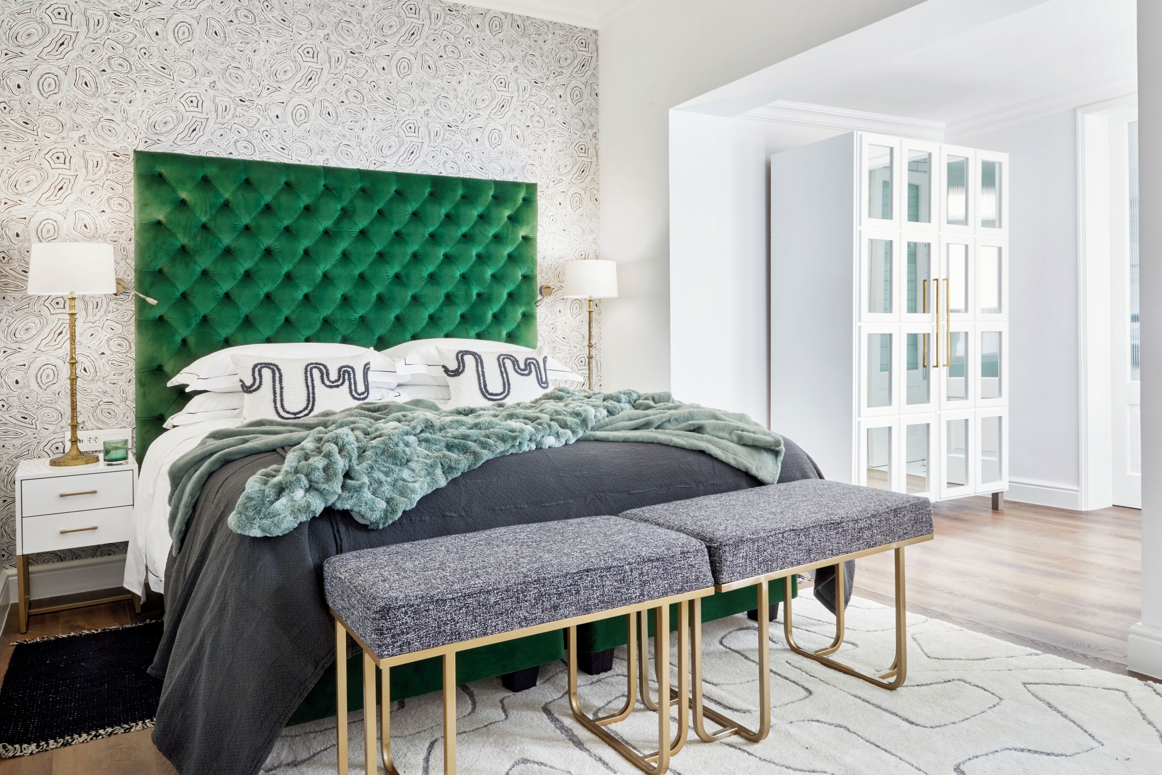 Un dormitorio moderno con un cabecero verde capitonado, un banco gris y un armario blanco con puertas de cristal.
