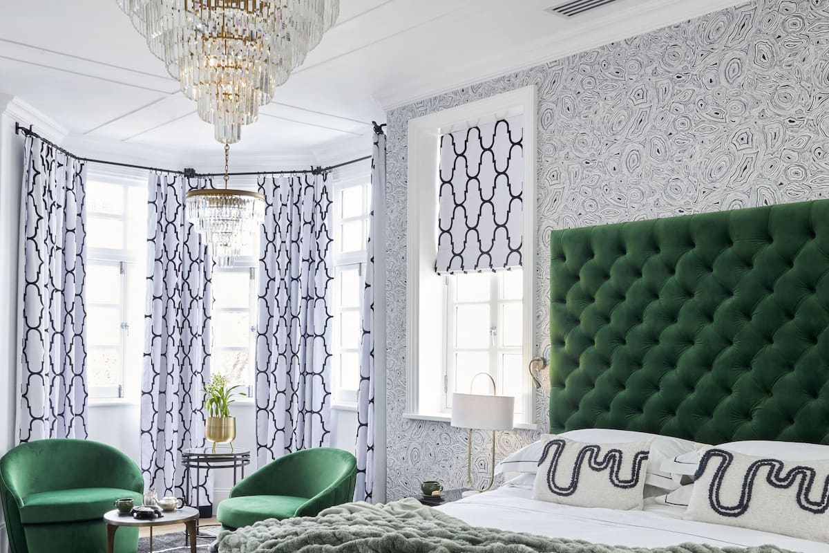 Een slaapkamer met een groen bekleden hoofdeinde, een groot bed met een grijs-witte gewatteerde spreideken, een bankje aan het voeteneind en een kroonluchter.