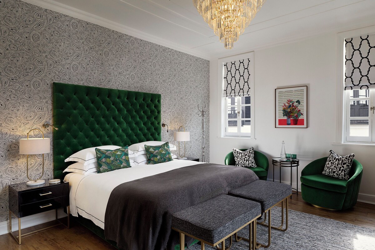 Een slaapkamer met een groen gestoffeerd hoofdeinde, een bed met witte lakens, twee nachtkastjes met lampen, een groene fauteuil, een klein rond tafeltje met een vaas, en een raam met patroon gordijnen.