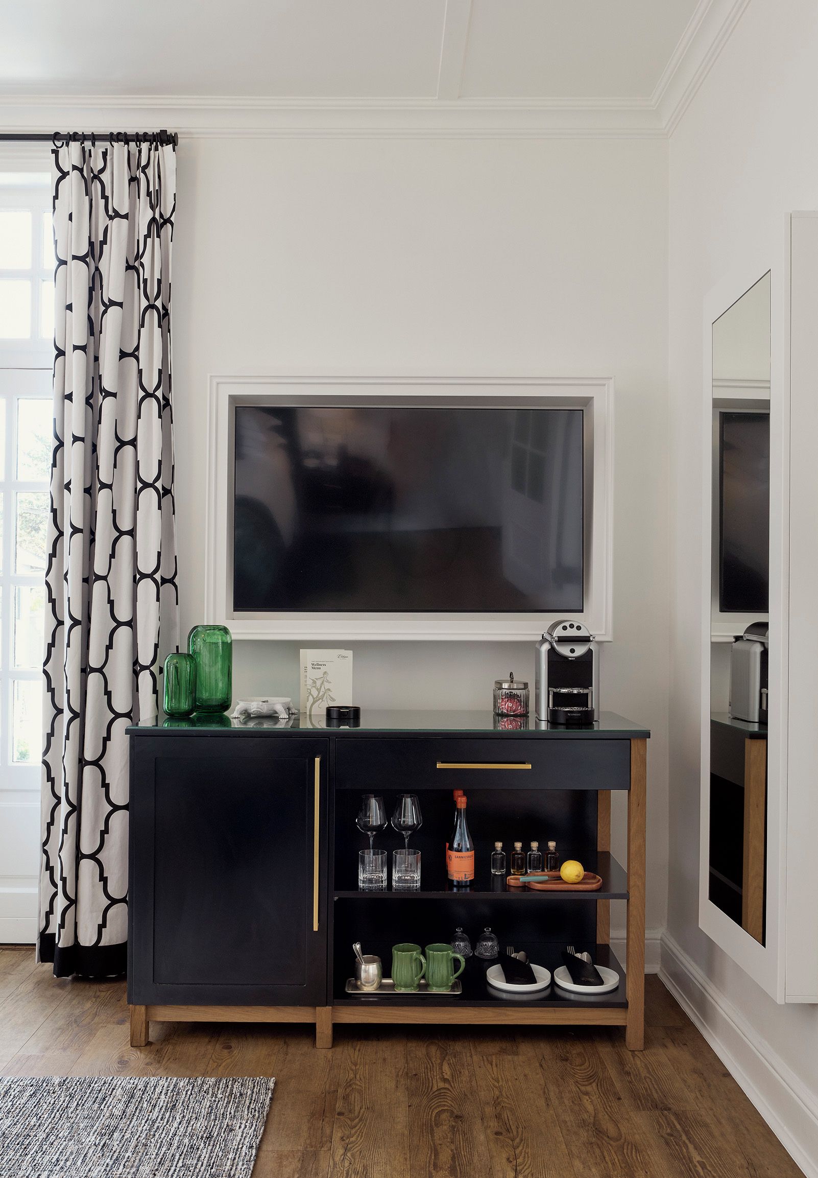 Ein modernes Wohnzimmer mit einem großen Flachbildfernseher an einer weißen Wand, einer schwarzen Unterhaltungseinheit darunter und einem gemusterten Vorhang am Fenster.