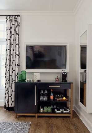 Een moderne woonkamer met een grote flatscreen televisie aan een witte muur, een zwart tv-meubel eronder en een patroon gordijn voor het raam.