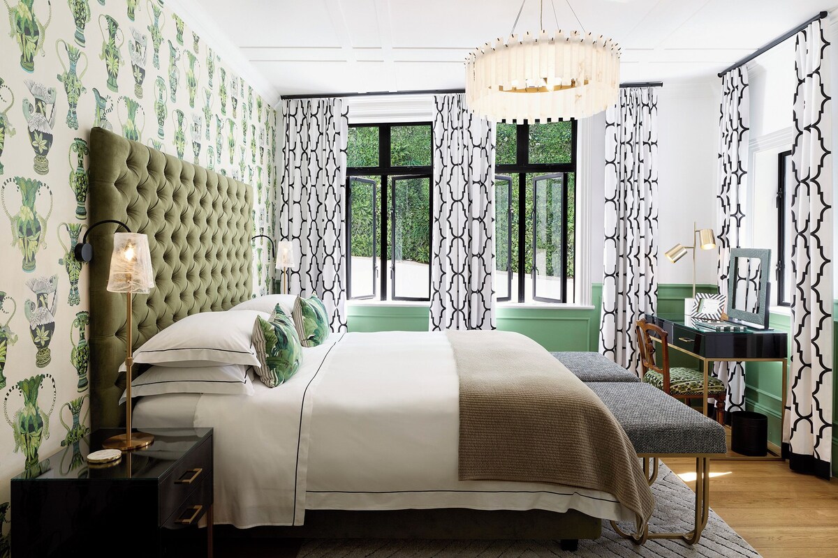 Een slaapkamer met een gestoffeerd hoofdeinde, een bed met witte beddengoed, een nachtkastje met een lamp en een bureau met een stoel.