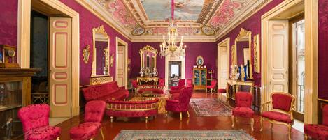 Una habitación lujosa con decoraciones ornamentales, incluyendo una gran lámpara de araña, sillones tapizados en rojo y una alfombra con un diseño detallado.