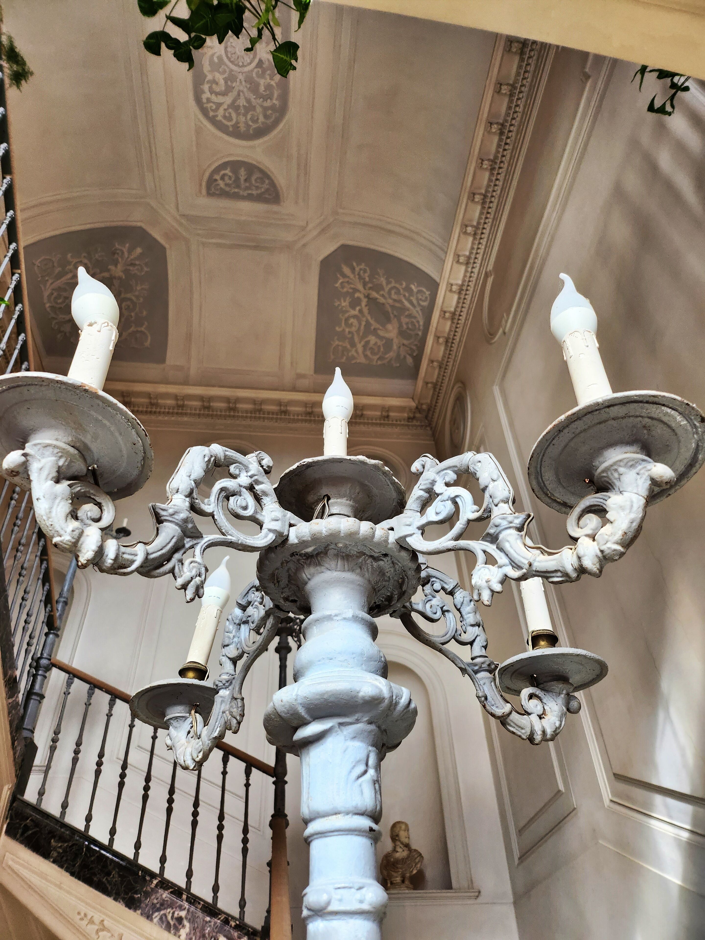 Un magnifique lustre, orné de multiples éléments rappelant des bougies, de motifs métalliques complexes et surplombant un plafond à l’architecture classique, agrémenté de moulures décoratives.