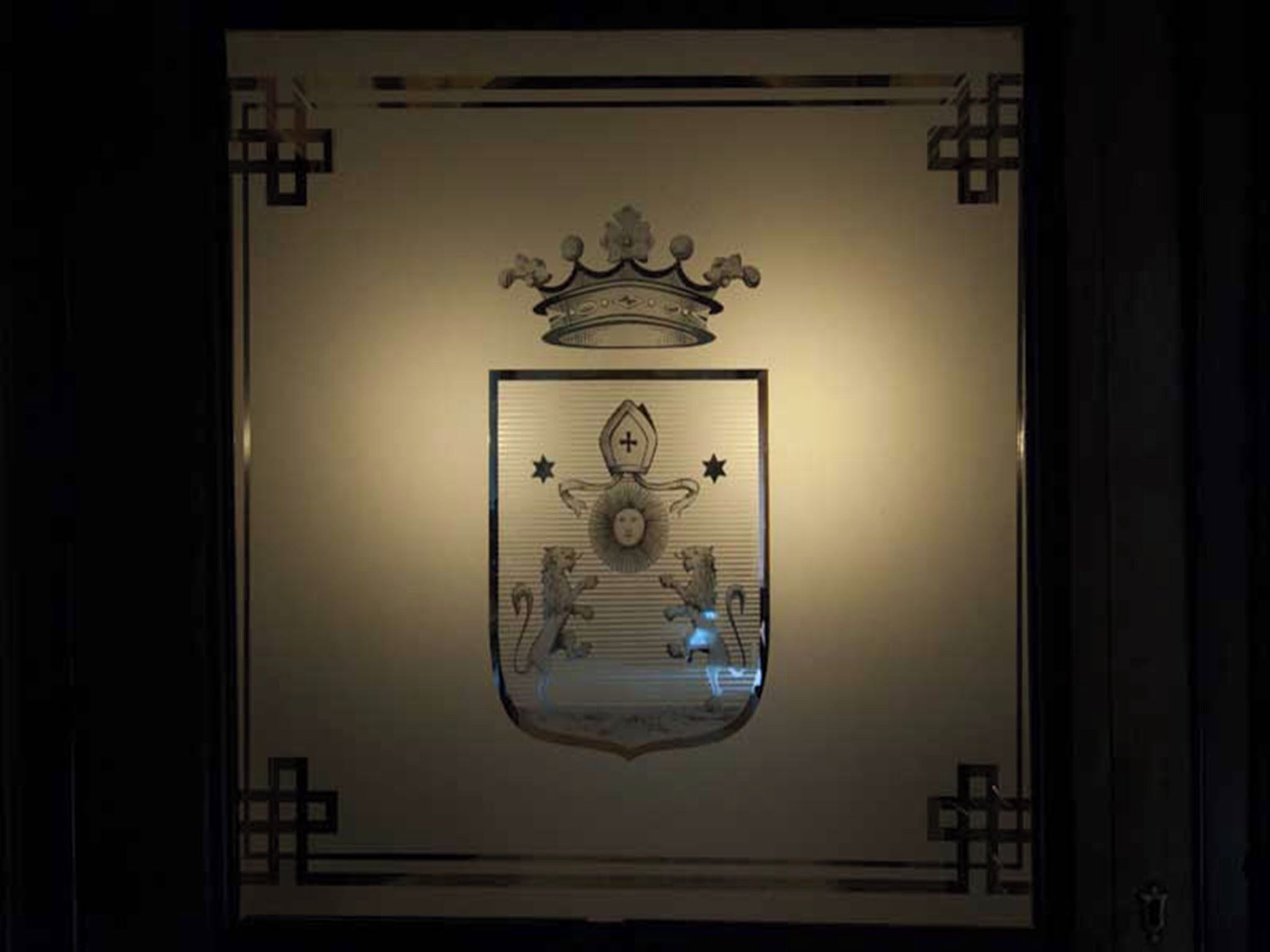 Um emblema emoldurado com uma coroa e um escudo, rodeado por elementos decorativos.
