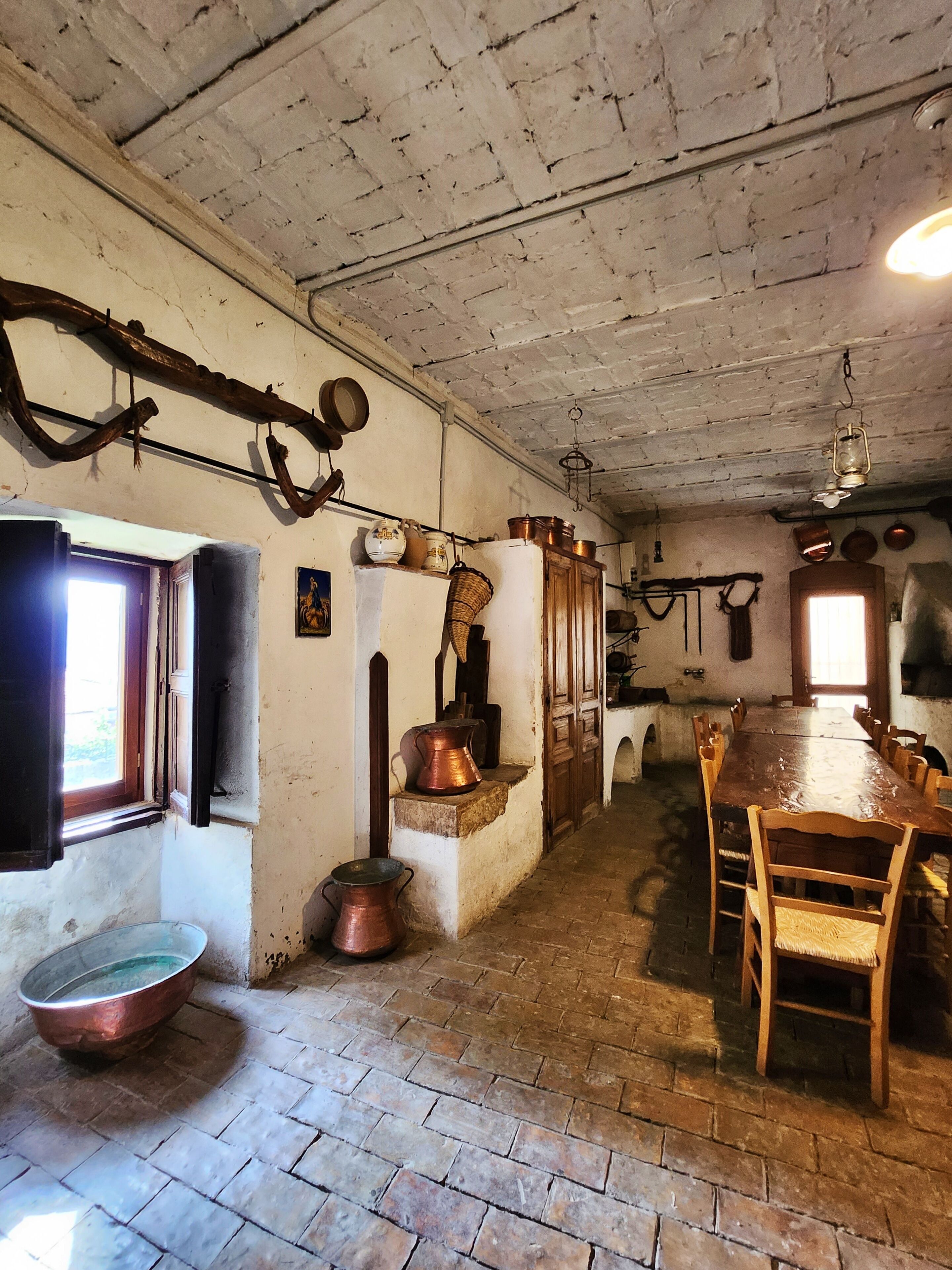 A rustic room with a long wooden table, chairs, and a brick floor. There are copper pots and pans hanging on the wall, and a window with curtains.