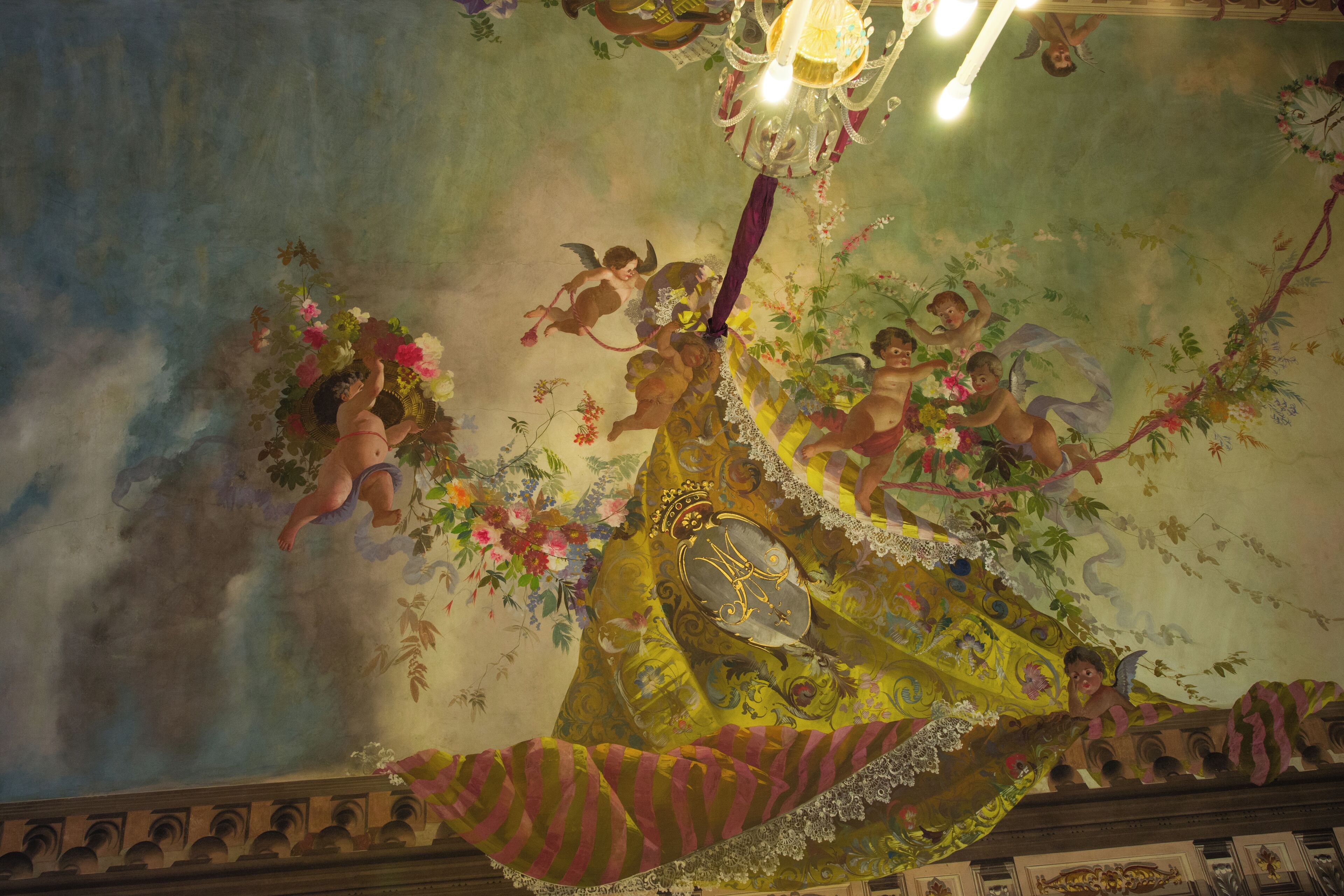Teto com um mural floral e com figuras de querubins, um candelabro e uma moldura estampada.